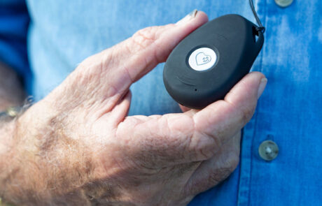 The image displays a gentleman's hand, holding a LiveLife personal alarm. The pendant alarm is black, matte with a round silver button in the center, that is the SOS button.