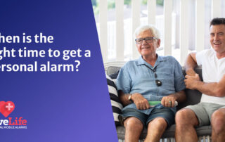 An elderly gentleman, wearing a personal alarm in black, sitting on the sofa in a home setting next to a younger man. Both are happy.