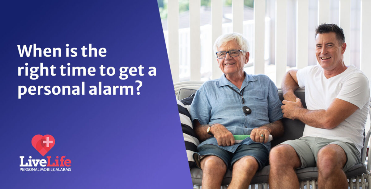 An elderly gentleman, wearing a personal alarm in black, sitting on the sofa in a home setting next to a younger man. Both are happy.