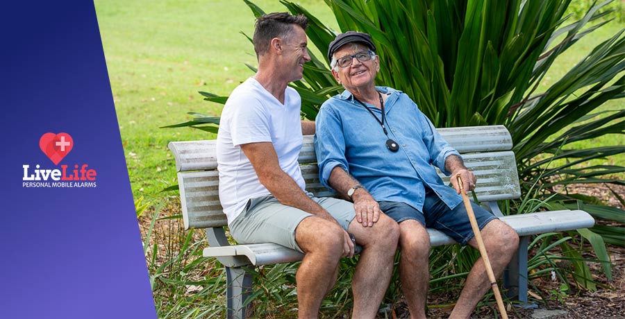 Senior wearing a livelife alarm, sitting on a park bench near a younger man.