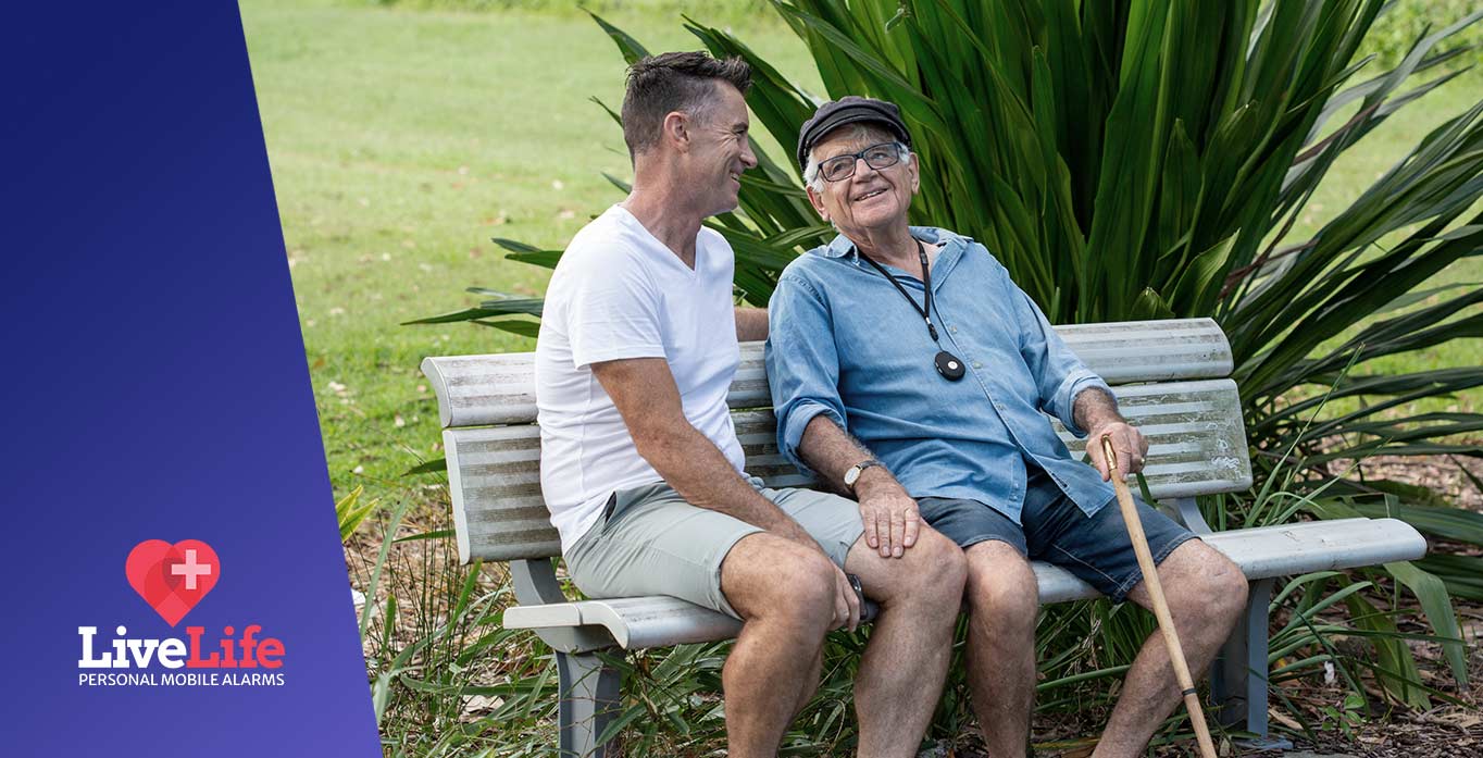 Senior elderly man sitting with son on bench. Man is wearing a Live Life personal medical alarm.